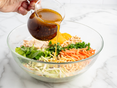 A hand pours a brown vinaigrette dressing from a glass pitcher onto a Chinese Chicken Salad in a clear bowl filled with lettuce, shredded vegetables, mandarin oranges, and crispy noodles—perfect for any Chinese Chicken Salad Recipe.