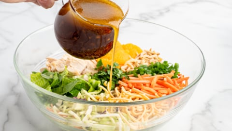 A hand pours a brown vinaigrette dressing from a glass pitcher onto a Chinese Chicken Salad in a clear bowl filled with lettuce, shredded vegetables, mandarin oranges, and crispy noodles&mdash;perfect for any Chinese Chicken Salad Recipe.