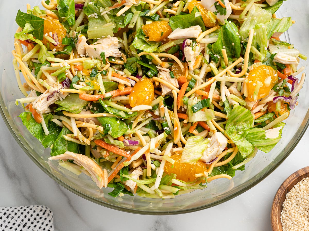 A close-up of a fresh Chinese Chicken Salad in a glass bowl, featuring shredded lettuce, carrots, cabbage, pieces of chicken, mandarin orange segments, and sesame seeds on a marble surface. Try this vibrant Chinese Chicken Salad Recipe!.