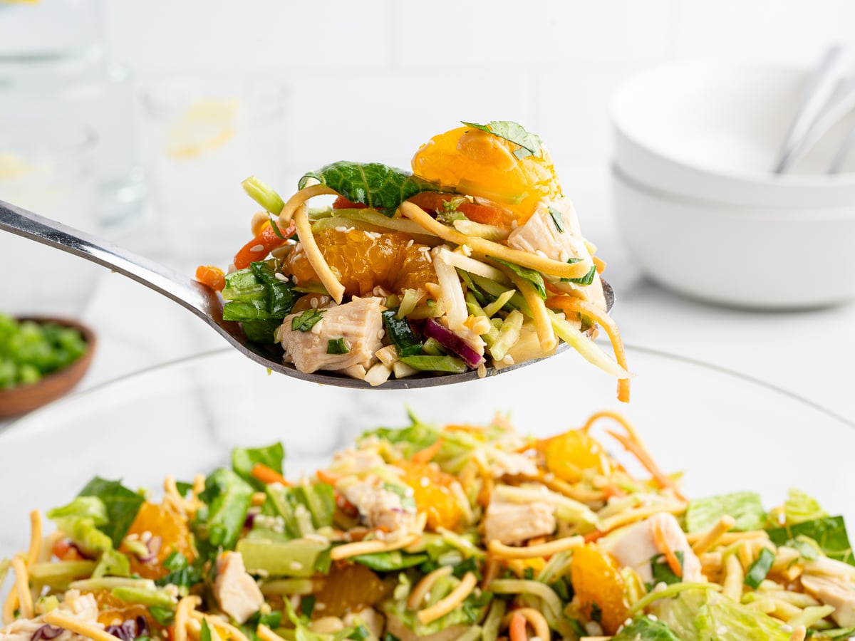 A close-up of a spoonful of Chinese Chicken Salad held above a bowl, featuring lettuce, shredded vegetables, mandarin orange segments, chunks of chicken, crispy noodles, and spinach. White bowls and utensils are blurred in the background.
