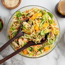 A glass bowl of colorful Chinese Chicken Salad with shredded chicken, lettuce, carrots, mandarin oranges, and green onions sits on a marble surface. Wooden salad servers rest nearby, along with dressing, sesame seeds, and platesโan inviting Chinese Chicken Salad Recipe.