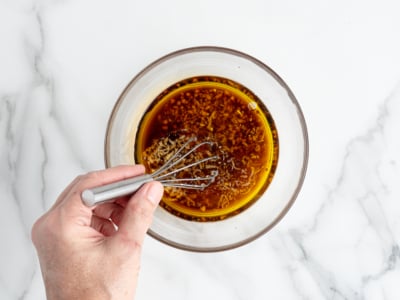 A hand using a small whisk to mix a brown sauce or marinade with visible minced garlic in a clear glass bowl on a white marble surface, perfect for drizzling over your favorite Chinese Chicken Salad Recipe.