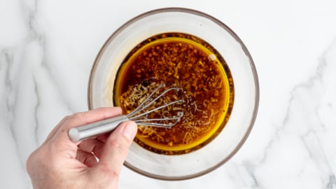 A hand using a small whisk to mix a brown sauce or marinade with visible minced garlic in a clear glass bowl on a white marble surface, perfect for drizzling over your favorite Chinese Chicken Salad Recipe.