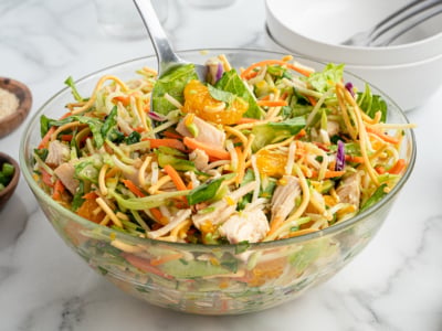 A glass bowl filled with Chinese Chicken Salad featuring shredded chicken, lettuce, mandarin orange slices, carrots, cabbage, and crunchy noodles, with a serving spoon inside. Plates and utensils are visible in the background.