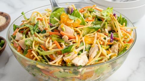 A glass bowl filled with Chinese Chicken Salad featuring shredded chicken, lettuce, mandarin orange slices, carrots, cabbage, and crunchy noodles, with a serving spoon inside. Plates and utensils are visible in the background.