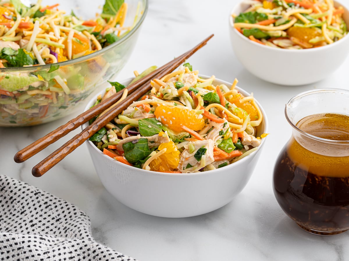 A white bowl filled with Chinese Chicken Salad featuring noodles, leafy greens, shredded carrots, and mandarin orange segments, with chopsticks resting on top. A glass bowl of salad, a smaller bowl, and a jar of dressing are nearby.