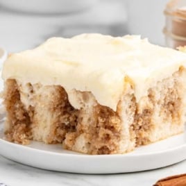 Slice of cinnamon roll poke cake on a white plate with cinnamon sticks in the background