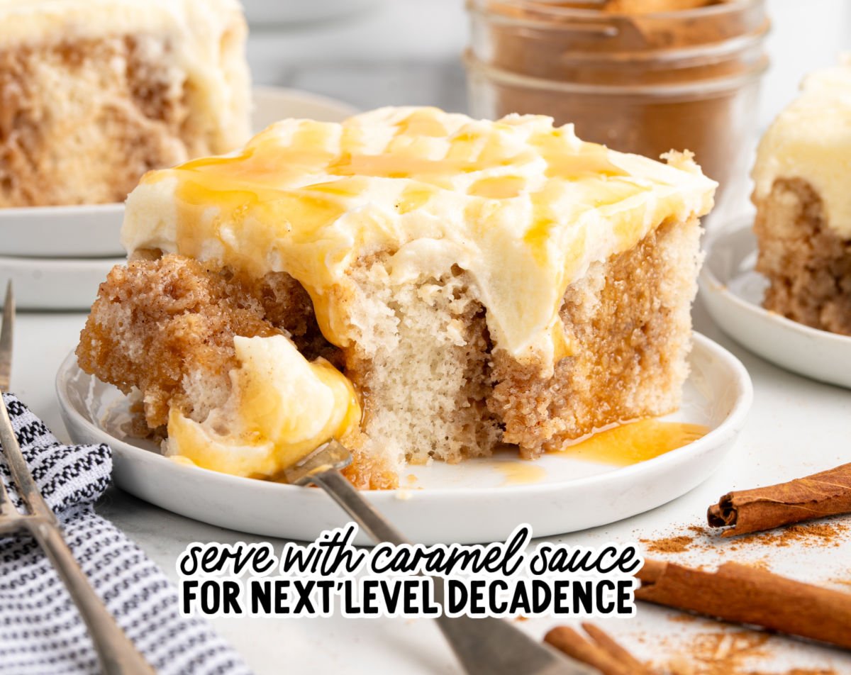Close-up slice of cinnamon roll poke cake topped with cream cheese frosting and drizzled with caramel sauce, served on a white plate with a fork, cinnamon sticks, and a jar of cinnamon in the background.