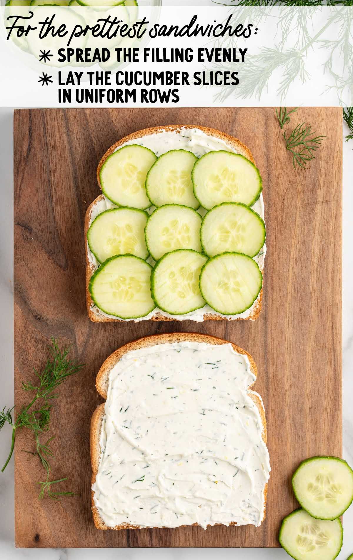 Overhead view of two slices of bread on a cutting board, one spread with cream cheese mixture and the other topped with cucumber slices.