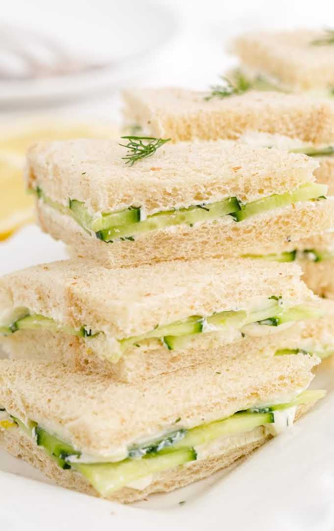 close up shot of cucumber sandwiches piled on top of each other on a plate garnished with fresh dill