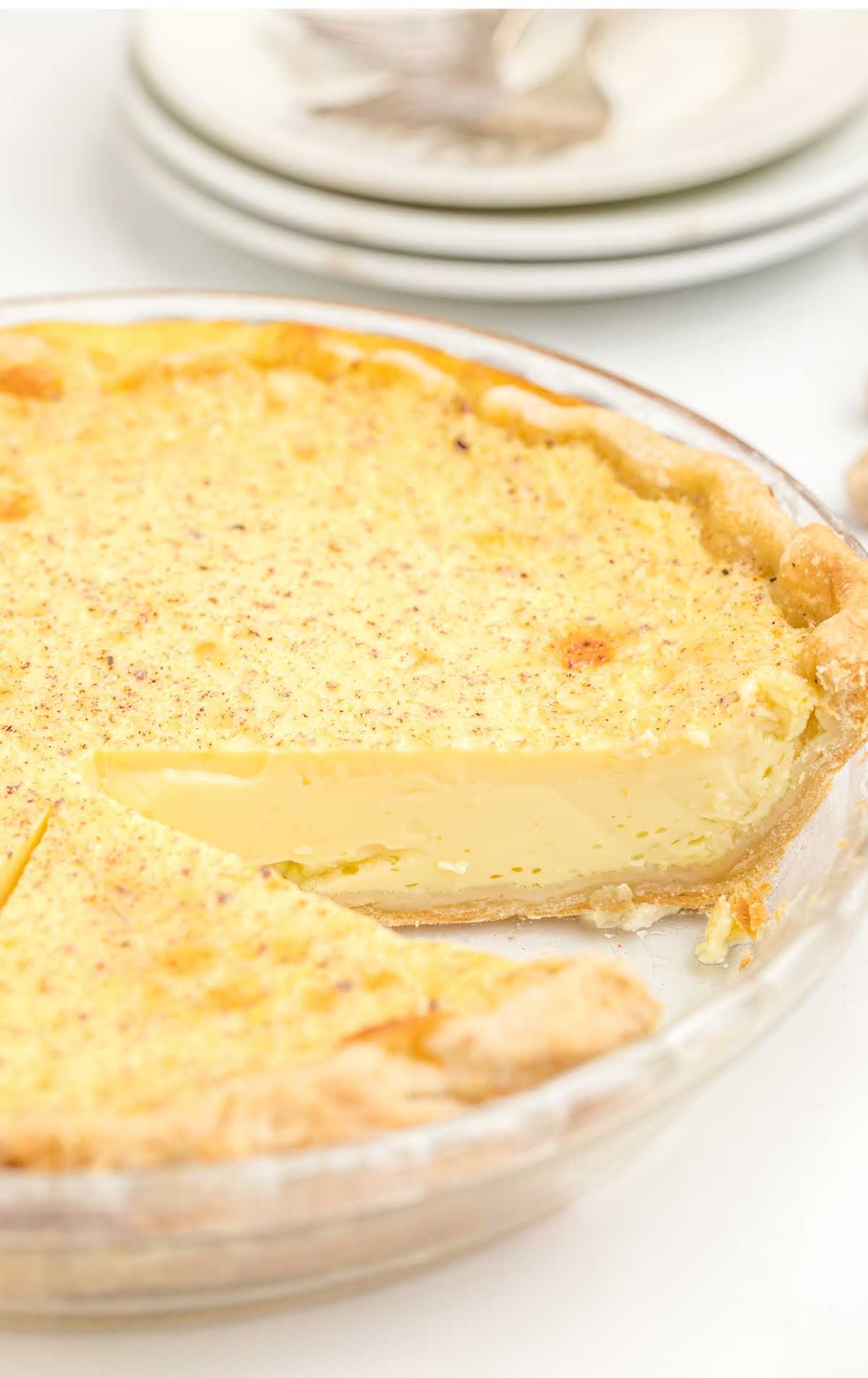 A freshly baked custard pie in a glass pie dish with a golden, flaky crust and a smooth, creamy filling. A slice has been removed, revealing the silky texture inside. A stack of white plates is visible in the background.