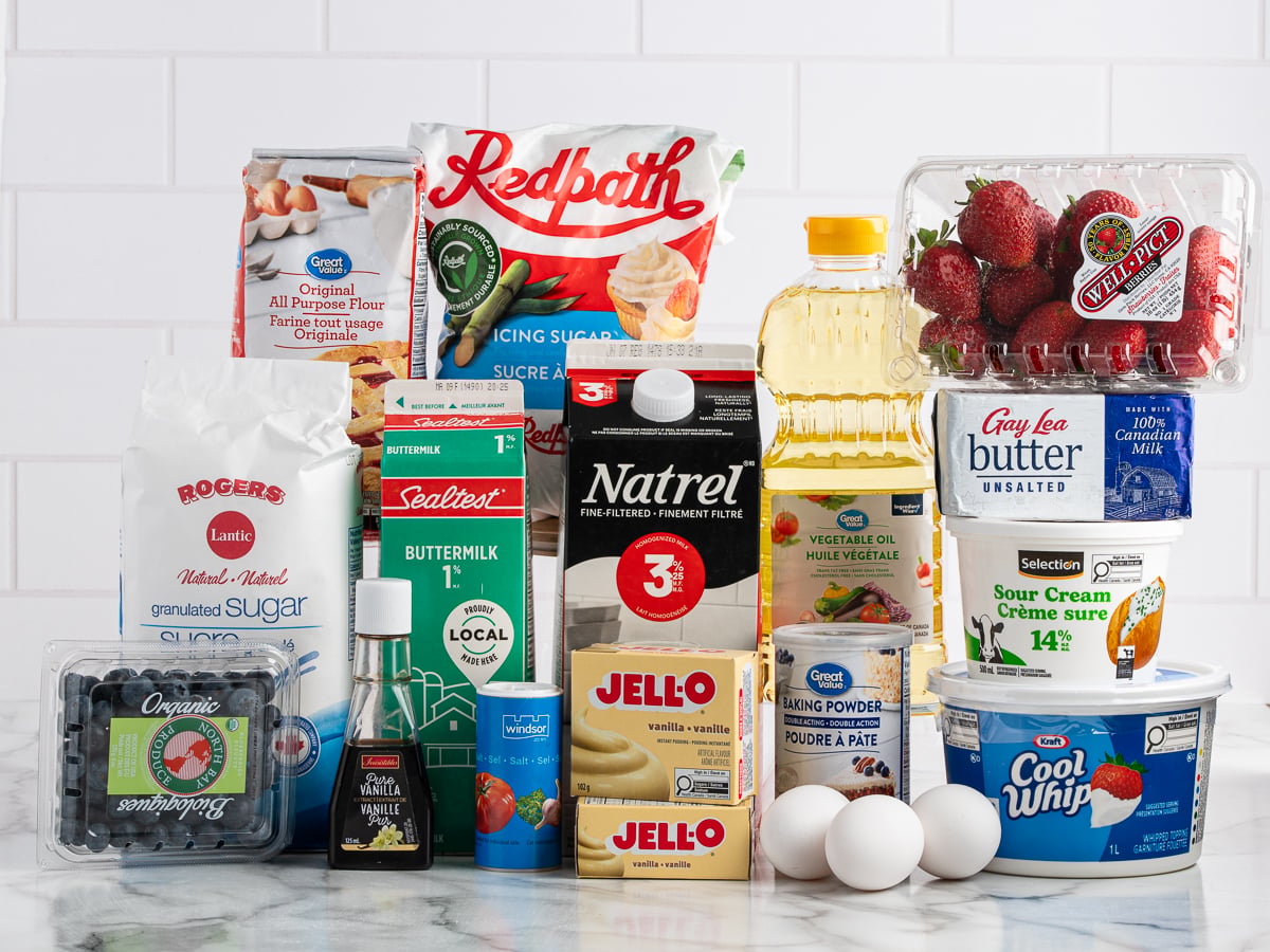 A variety of baking ingredients arranged on a white counter, ready to make an American Flag Cake, including flour, sugar, buttermilk, vanilla, milk, Jello pudding, baking powder, oil, butter, strawberries, blueberries, sour cream, Cool Whip, and two eggs.