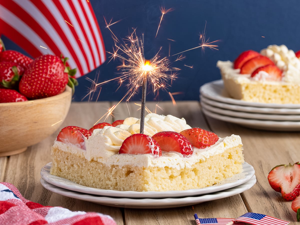 A slice of vanilla cake topped with whipped cream and strawberries sits on a plate, sparkler aglow. An American Flag Cake and a bowl of strawberries add festive flair in the background, perfect for your next American Flag Cake recipe celebration.