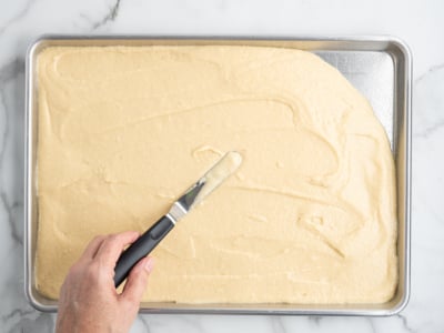 A hand is using an offset spatula to evenly spread batter for an American Flag Cake onto a rectangular metal baking sheet on a white marble surface.