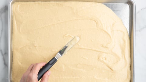 A hand is using an offset spatula to evenly spread batter for an American Flag Cake onto a rectangular metal baking sheet on a white marble surface.