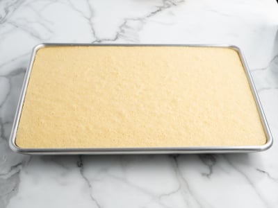 A rectangular sheet pan filled with a layer of plain yellow cake, ready to be transformed into a festive American Flag Cake on a white marble surface.
