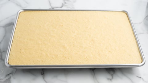 A rectangular sheet pan filled with a layer of plain yellow cake, ready to be transformed into a festive American Flag Cake on a white marble surface.