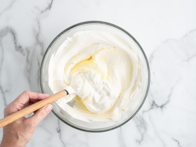 A hand stirs a creamy white mixture with a spatula in a glass bowl on a white marble surface, preparing the base for an American Flag Cake Recipe.