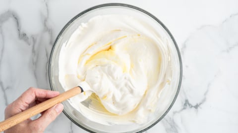 A hand stirs a creamy white mixture with a spatula in a glass bowl on a white marble surface, preparing the base for an American Flag Cake Recipe.