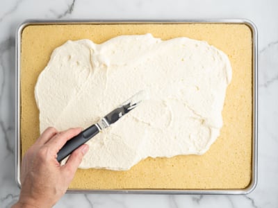 A hand uses an offset spatula to spread white frosting over a rectangular sheet cake on a metal baking tray, preparing the base for an American Flag Cake, set on a marble surface.