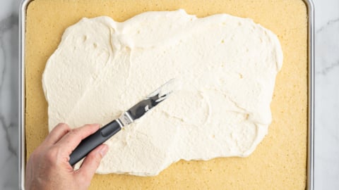 A hand uses an offset spatula to spread white frosting over a rectangular sheet cake on a metal baking tray, preparing the base for an American Flag Cake, set on a marble surface.