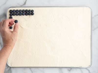 A hand places blueberries in neat rows on the top left corner of a large rectangular tray filled with creamy white dessert, set on a marble surface—perfect for an American Flag Cake.