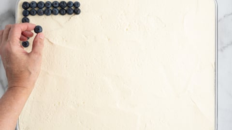 A hand places blueberries in neat rows on the top left corner of a large rectangular tray filled with creamy white dessert, set on a marble surface—perfect for an American Flag Cake.