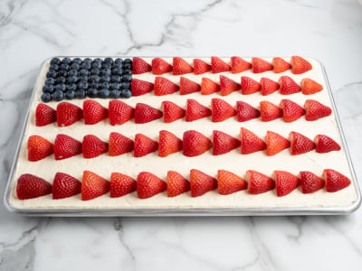 An American Flag Cake sits on a marble surface, decorated with blueberries for the blue corner and rows of halved strawberries as red stripes atop white frosting—a perfect treat for your next American Flag Cake Recipe celebration.