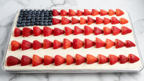 An American Flag Cake sits on a marble surface, decorated with blueberries for the blue corner and rows of halved strawberries as red stripes atop white frosting—a perfect treat for your next American Flag Cake Recipe celebration.