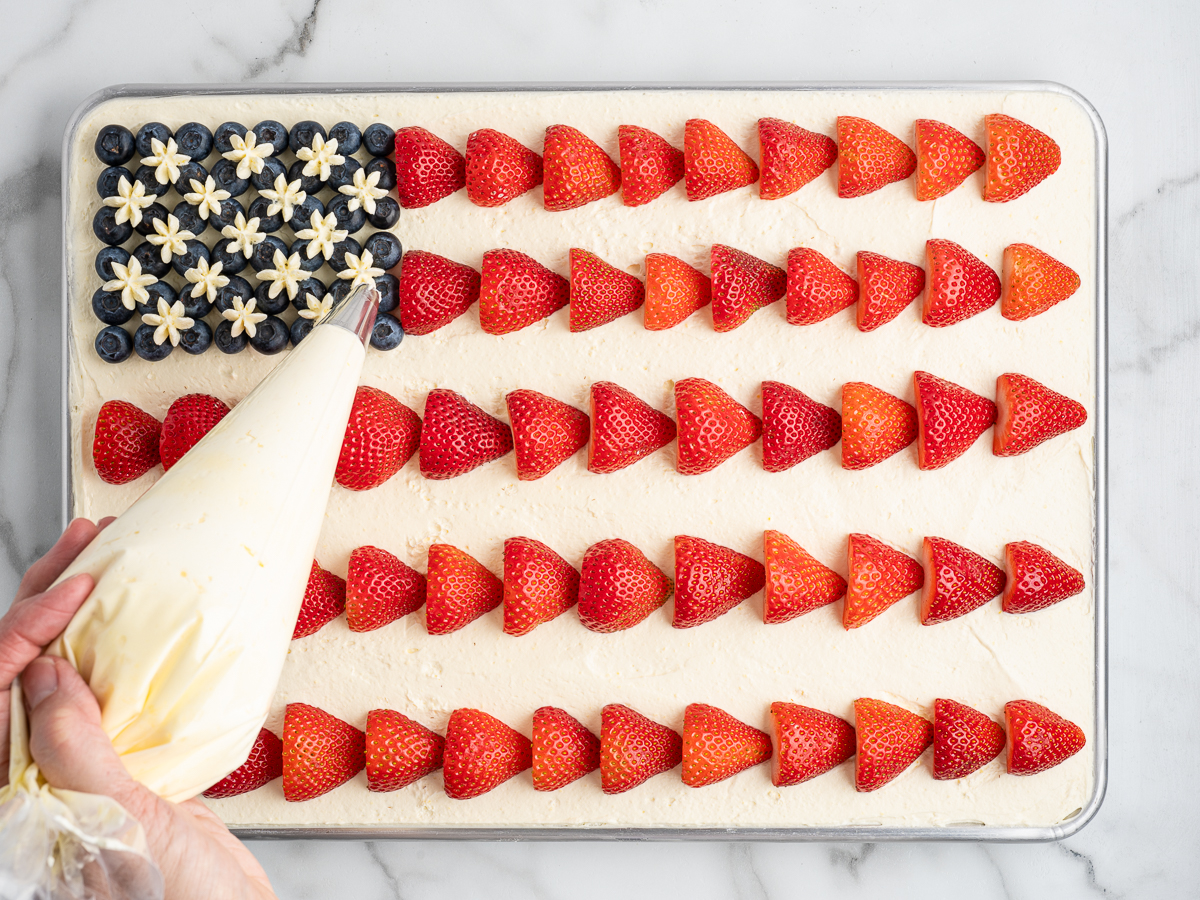A hand pipes white frosting stars onto blueberries in the top left corner of an American Flag Cake decorated with rows of strawberry halves, resembling the stars and stripes.