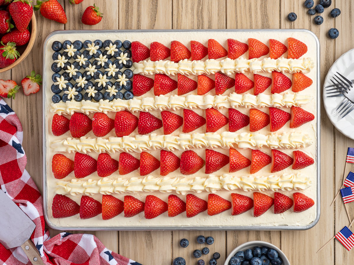 An American Flag Cake decorated with strawberries, piped white frosting, and blueberries sits on a wooden table with fresh berries and utensils nearby&mdash;perfect for any patriotic celebration. Try this classic American Flag Cake recipe today!.