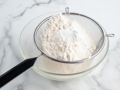A metal sifter holding flour and baking powder is positioned over a clear glass mixing bowl on a white marble surface, prepping ingredients for an American Flag Cake Recipe.