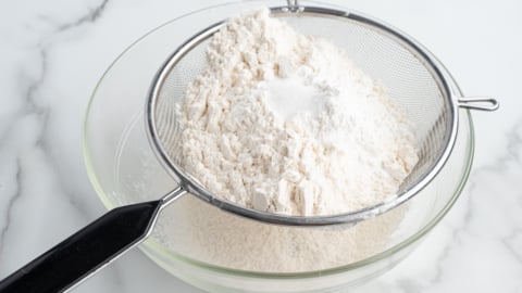 A metal sifter holding flour and baking powder is positioned over a clear glass mixing bowl on a white marble surface, prepping ingredients for an American Flag Cake Recipe.