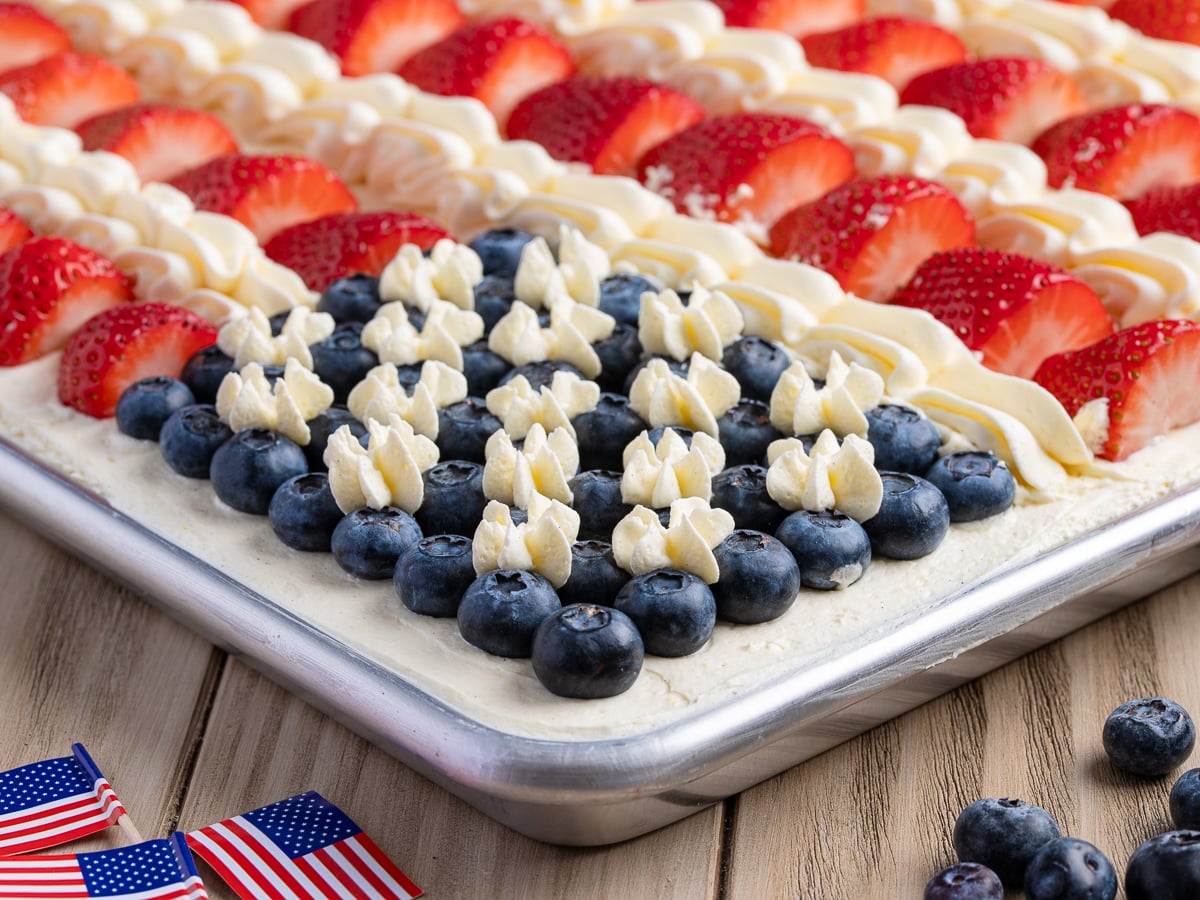 A classic American Flag Cake decorated with sliced strawberries, blueberries, and white frosting stars. Small American flag picks rest on the wooden table next to this patriotic dessert—a perfect treat for any celebration!.
