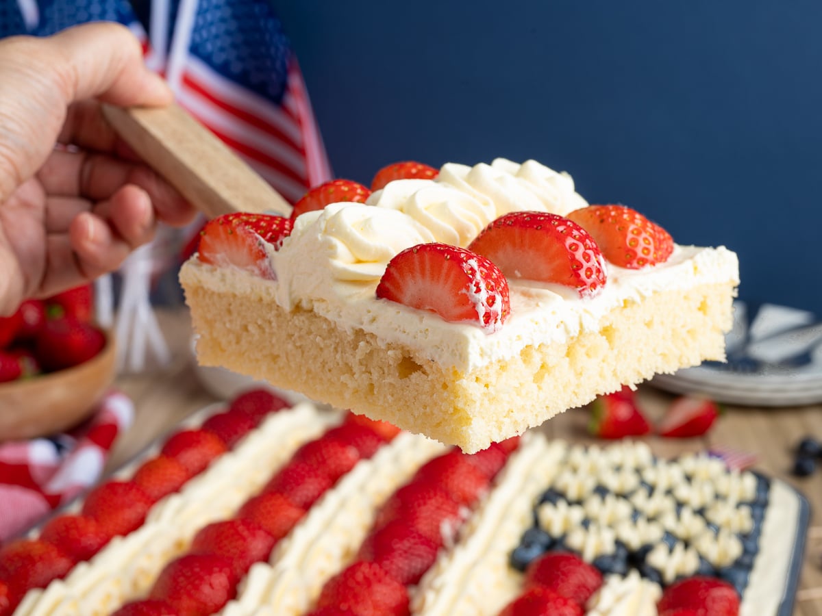 A hand holds a slice of cake topped with whipped cream and strawberries. In the background, an American Flag Cake decorated with strawberries, blueberries, and small flags is visible.