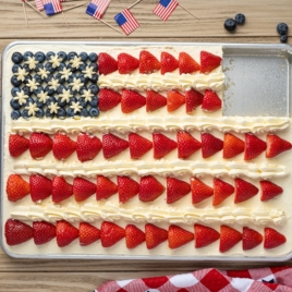 An American Flag Cake decorated with blueberries and star-shaped icing for the stars, and rows of sliced strawberries for the stripes. A corner piece is missing. Small US flags and blueberries are nearby on the table.
