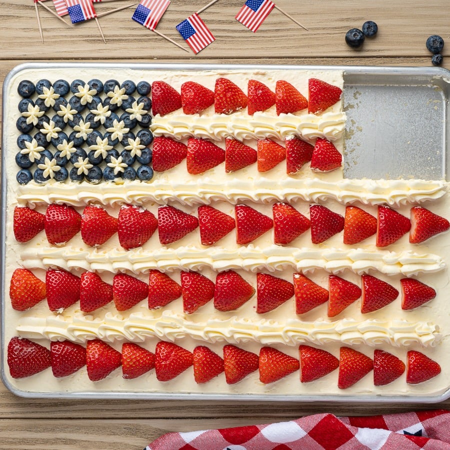 An American Flag Cake decorated with blueberries and star-shaped icing for the stars, and rows of sliced strawberries for the stripes. A corner piece is missing. Small US flags and blueberries are nearby on the table.