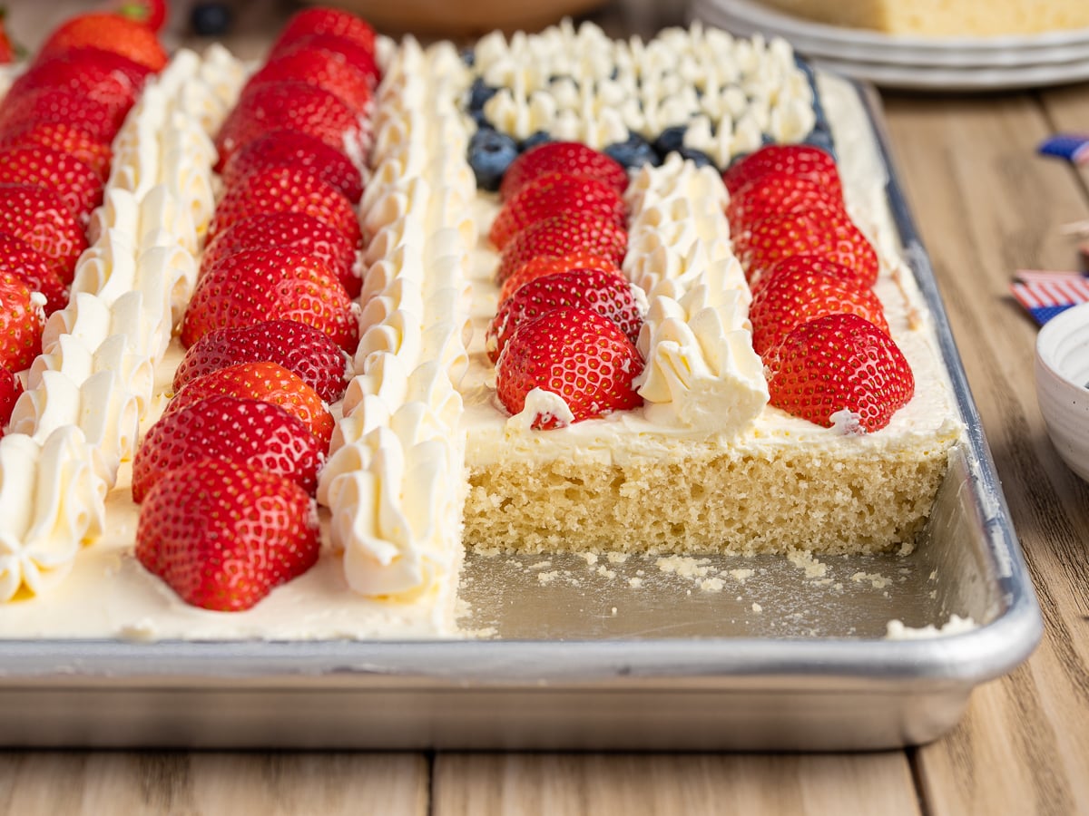 Sheet cake decorated like an American Flag Cake with rows of strawberries, whipped cream, and blueberries on white frosting. A rectangular slice has been removed from the lower right corner, revealing the fluffy cake inside.