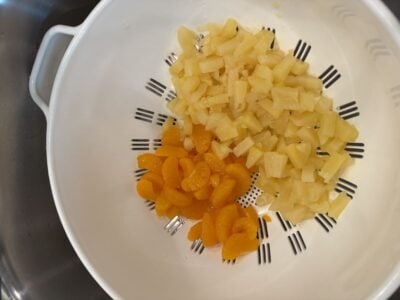 Chopped pineapple and sliced mandarin oranges, perfect for Ambrosia Salad, are draining in a white colander with black lines, placed over a sink.