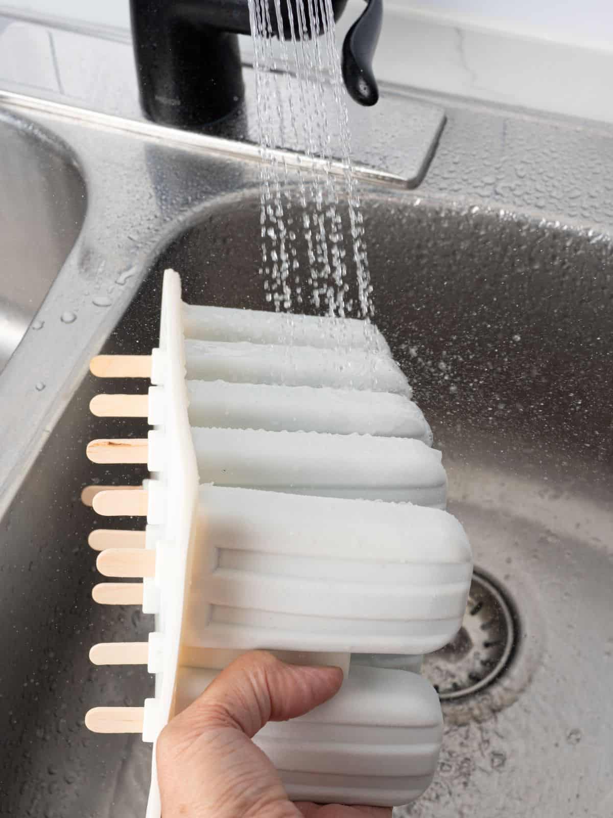 A hand holds a popsicle mold under running water in a kitchen sink, loosening the frozen treats for easy removalโperfect for enjoying homemade Fudgesicles from your favorite Fudgesicles recipe.
