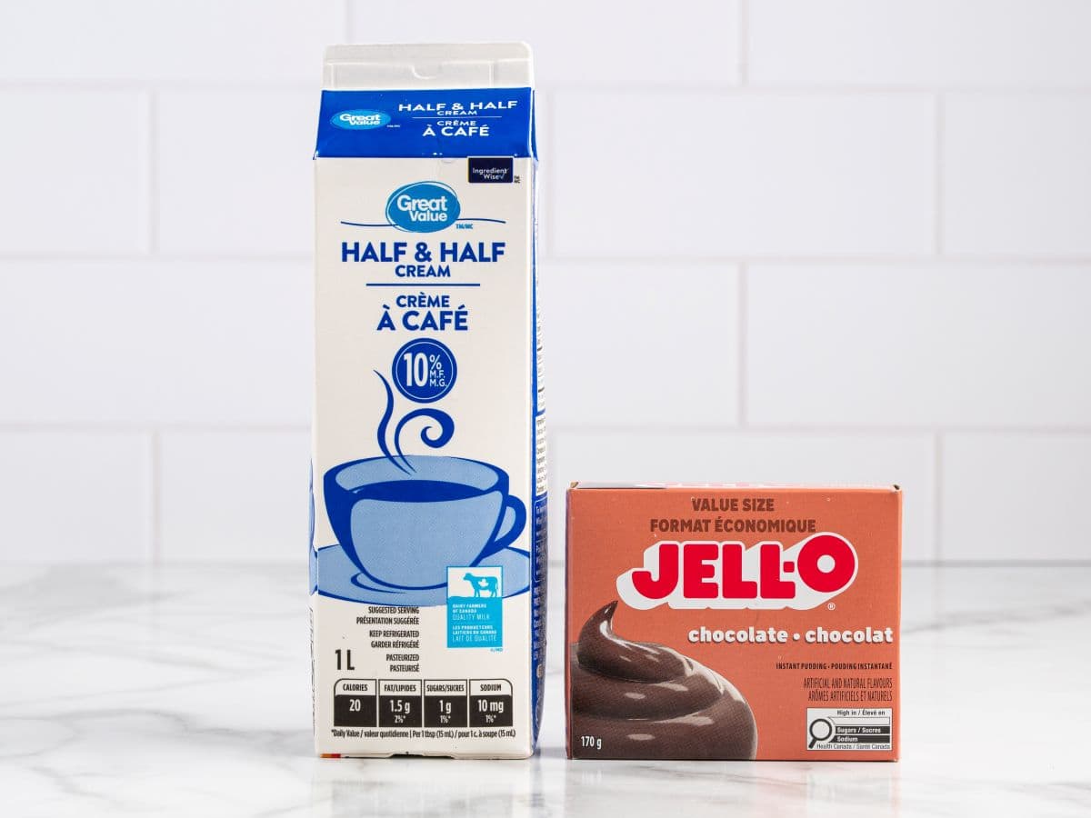 A carton of Great Value Half & Half Cream sits next to a box of Jell-O chocolate pudding on a white countertop with a white tile backgroundโperfect ingredients for making a creamy homemade Fudgesicles Recipe.