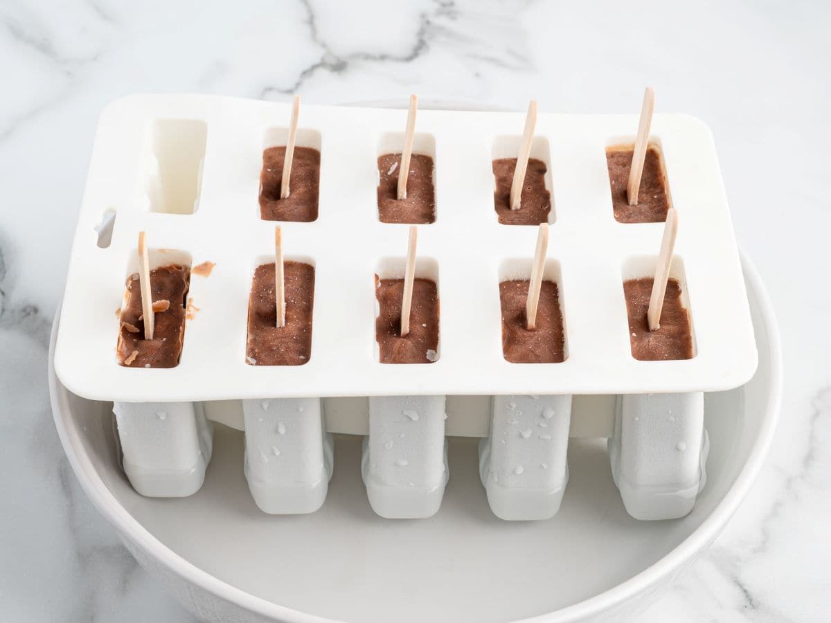 A white popsicle mold filled with a rich chocolate mixture and wooden sticks sits on a white plate, with some drips and droplets visible on the mold and plateโperfect for making your favorite homemade Fudgesicles Recipe.