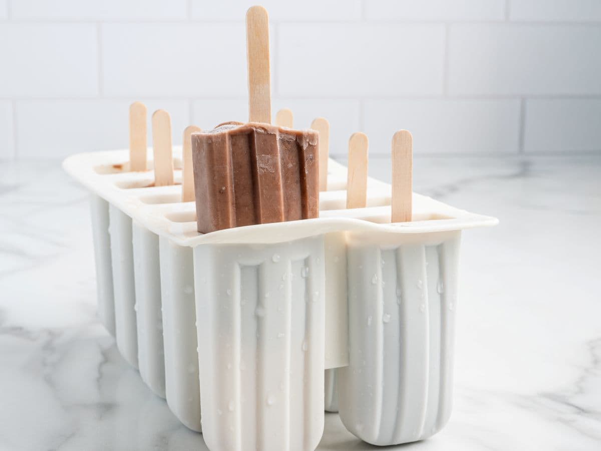A white popsicle mold tray holds several popsicles, including a rich chocolate Fudgesicle raised above the others. The tray sits on a marble countertop with a tiled wall in the background, tempting anyone looking for an easy homemade Fudgesicles recipe.