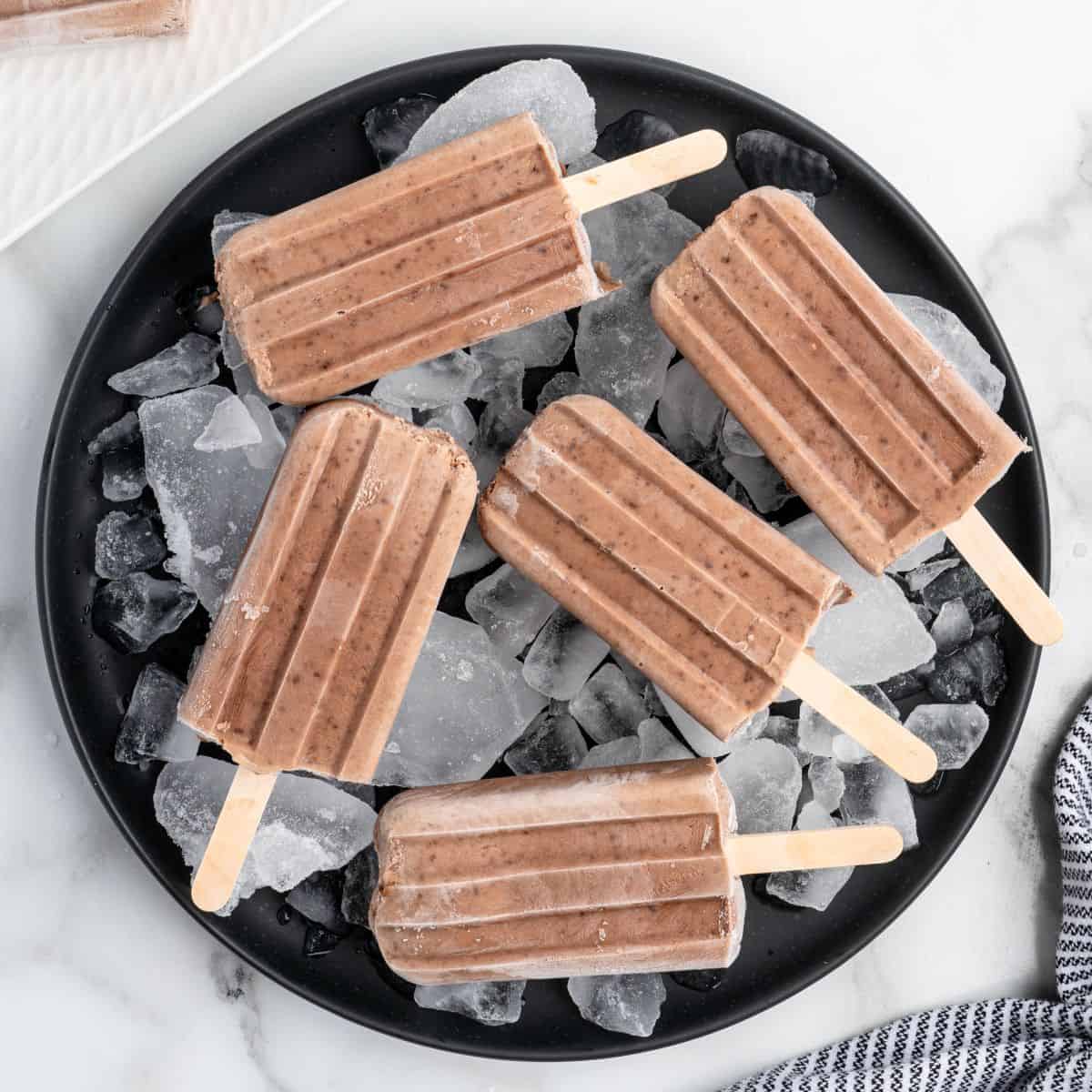 Five chocolate Fudgesicles on wooden sticks are arranged on a round black plate filled with ice cubes, set on a white marble surface. A portion of a striped cloth is visible in the corner, perfectly complementing this delightful homemade Fudgesicles recipe scene.