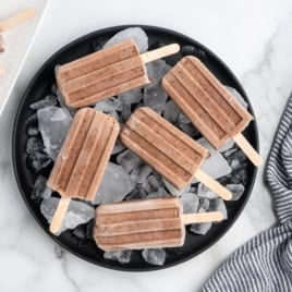 Fudgesicles arranged on a black plate filled with ice cubes, set on a white marble surface next to a striped cloth, make for an irresistible homemade Fudgesicles Recipe perfect for cooling down on warm days.