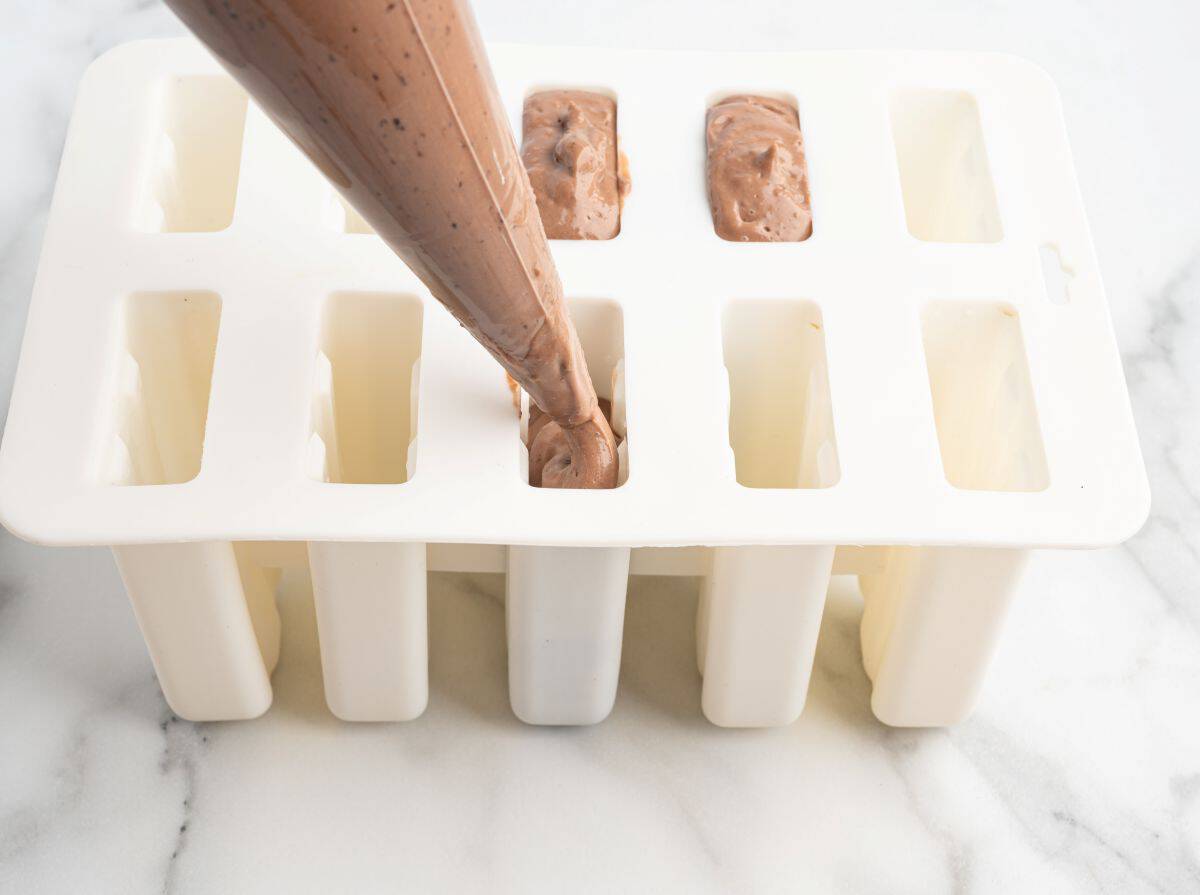 A piping bag fills a white popsicle mold with a rich chocolate mixture, perfect for a homemade Fudgesicles Recipe. Some molds are already filled, while others remain empty on a light marble surface, eagerly awaiting their turn to become delicious Fudgesicles.