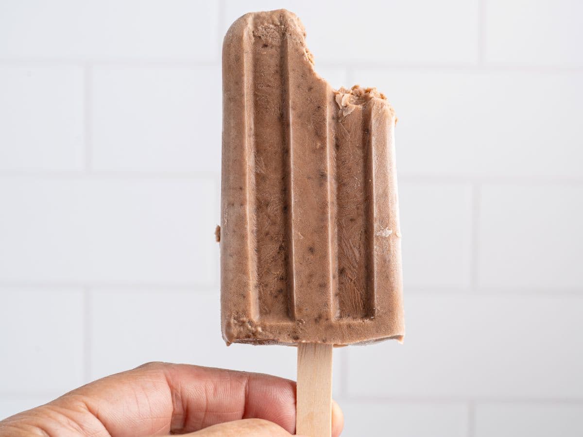 A hand holds a chocolate Fudgesicle with a bite taken out of the top against a white tiled background, perfectly capturing the irresistible appeal of this classic Fudgesicles recipe.