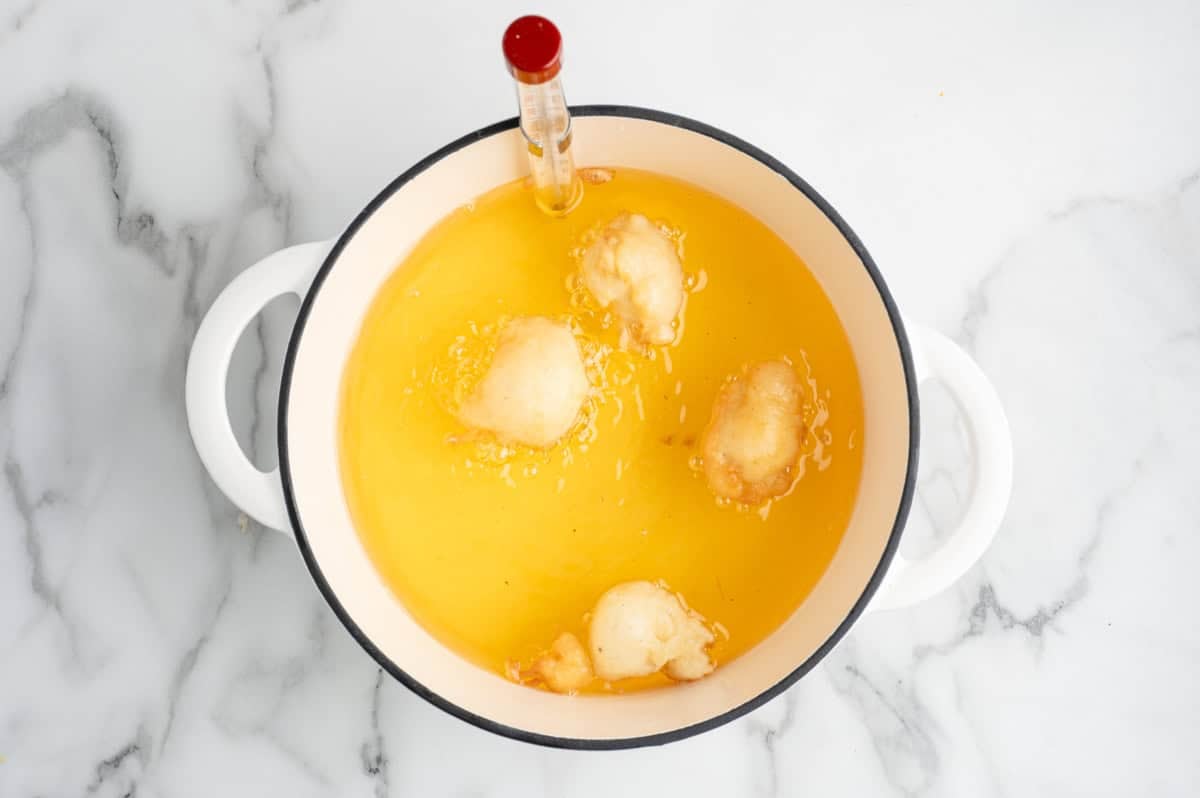 A white pot with oil and four pieces of dough frying inside, perfect for a Funnel Cake Bites Recipe, with a cooking thermometer clipped to the side, sits on a white marble surface.