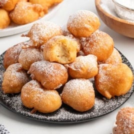 A black plate piled with golden, round Funnel Cake Bites dusted with powdered sugar. One fritter at the top is bitten in half, revealing a soft, airy interior. Other dishes with funnel cake bites and powdered sugar are in the background.