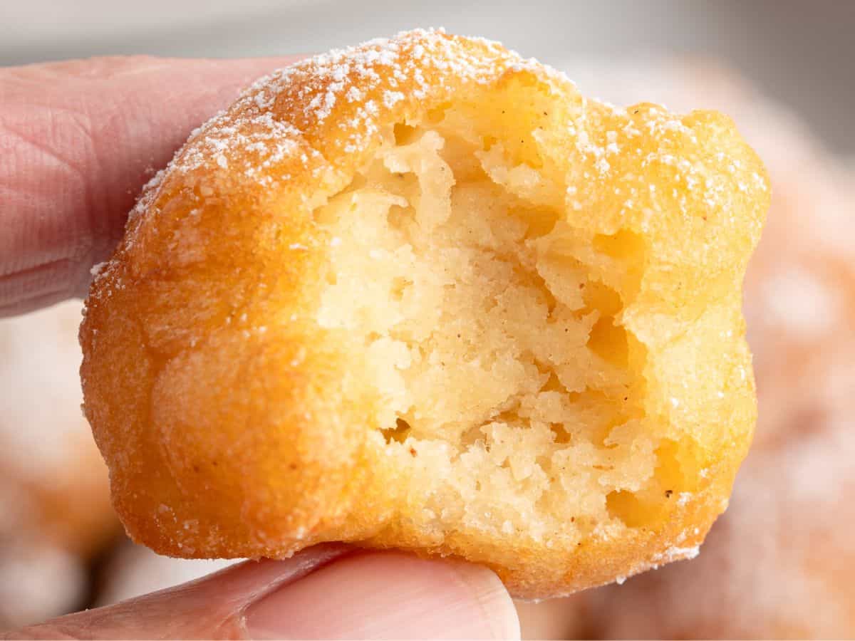 A close-up of a hand holding a golden, fluffy Funnel Cake Bite with a bite taken out, showing its soft, airy interior and topped with a dusting of powdered sugar.